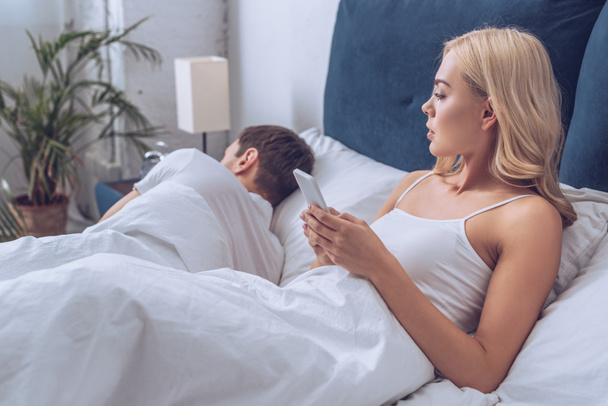 jealous young woman using smartphone and looking at boyfriend sleeping in bed, distrust concept - Foto, Imagem