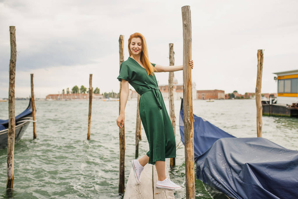 Young beautiful girl with red hair in green clothes stands near the water overlooking beautiful Venice - Фото, зображення