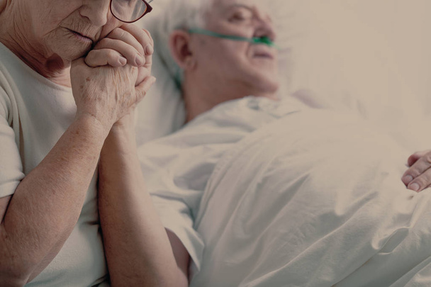 Worried senior woman holding hand of her sick husband with lupus lying in hospital bed, photo with copy space - 写真・画像