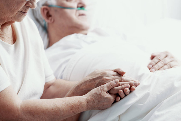 Senior man with lung cancer laying in hospice bed supporting by his elderly wife, photo with copy space - Фото, изображение