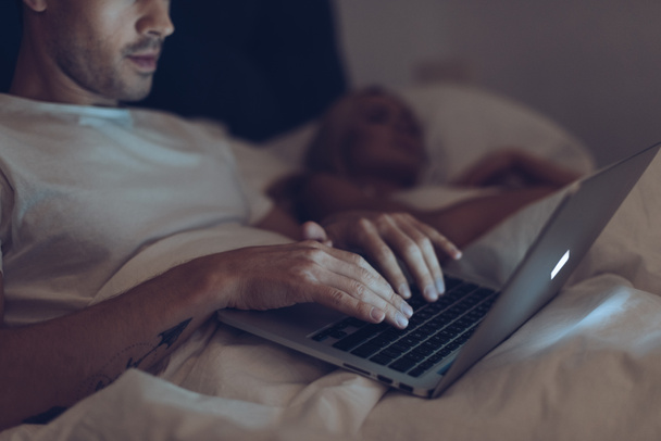 Schnappschuss von verdächtigem jungen Mann mit Laptop, während Frau nachts im Bett schläft  - Foto, Bild
