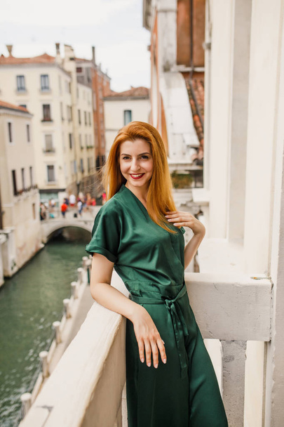 A young beautiful girl with red hair in green clothes is standing on a balcony overlooking beautiful Venice - Фото, зображення