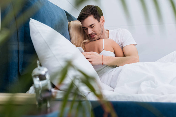 selective focus of smiling young man using smartphone while lying with girlfriend in bed - Fotoğraf, Görsel