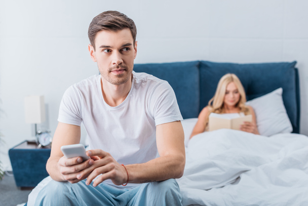 Junger Mann hält Smartphone in der Hand und schaut weg, während Freundin im Bett Buch liest - Foto, Bild