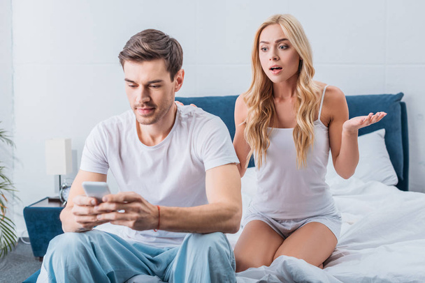 όμορφη σοκαρισμένος γυναίκα ψάχνει άντρα που κάθεται στο κρεβάτι και χρησιμοποιώντας το smartphone, δυσπιστία έννοια - Φωτογραφία, εικόνα