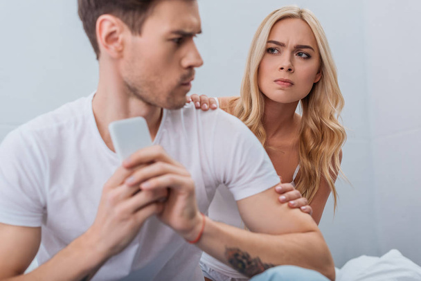angry young woman looking at boyfriend using smartphone in bedroom, relationship problem concept - Valokuva, kuva