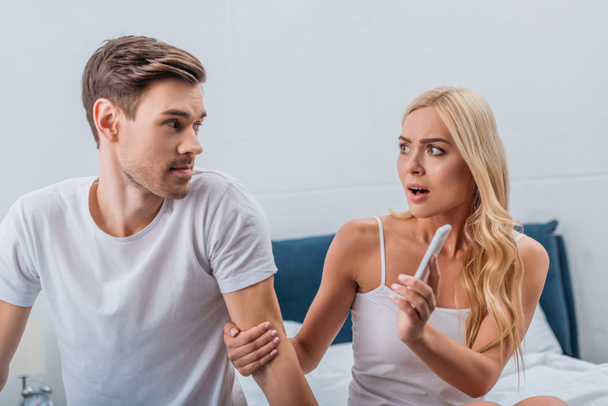 angry woman showing smartphone to young husband sitting on bed, relationship problem concept - Photo, Image