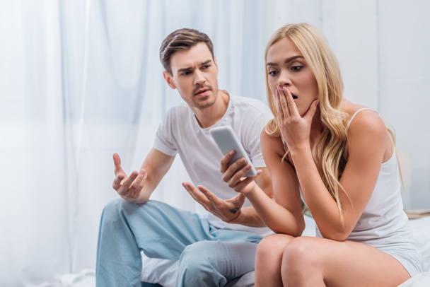 Emotionaler junger Mann gestikuliert mit den Händen und schaut schockierte Freundin mit Smartphone im Bett an - Foto, Bild