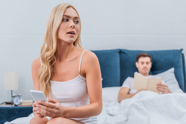 young woman using smartphone and looking away while husband reading book on bed  - Foto, Bild