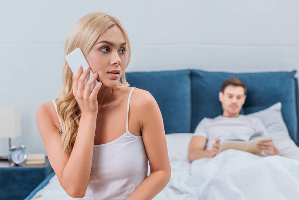 jonge vrouw praten door smartphone en op zoek weg terwijl echtgenoot leesboek op bed  - Foto, afbeelding