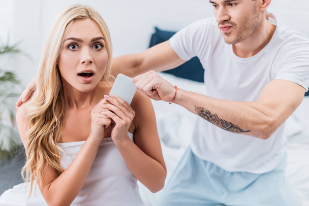 jealous man grabbing smartphone from scared girlfriend looking at camera in bedroom - Photo, Image