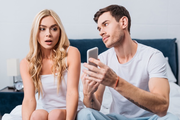 jealous man showing smartphone to confused girlfriend sitting on bed, distrust concept  - Foto, Imagen
