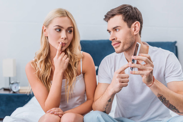 jealous man pointing at smartphone to confused girlfriend sitting on bed, distrust concept  - Photo, Image