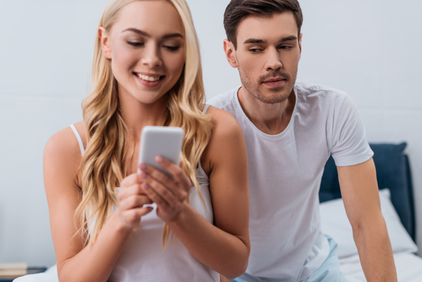 jealous man looking at beautiful smiling wife using smartphone in bedroom, mistrust concept  - Photo, Image