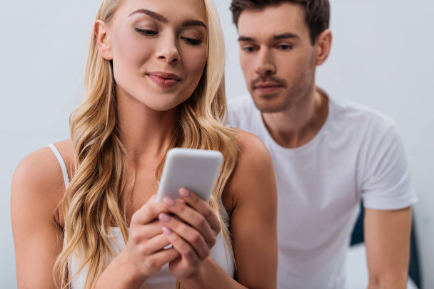 man looking at beautiful smiling wife using smartphone in bedroom, mistrust concept  - Photo, Image