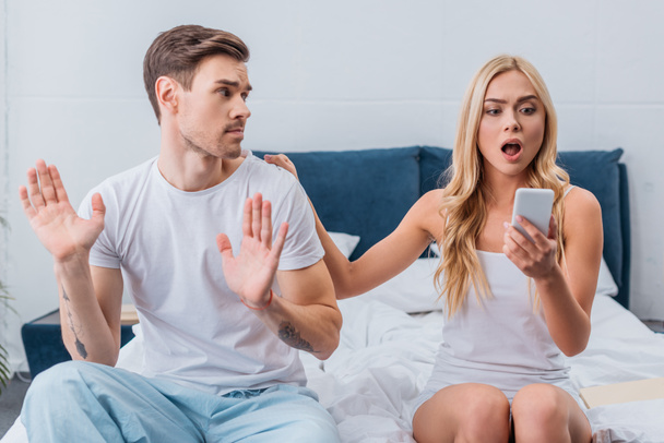 shocked young woman using smartphone while sitting with boyfriend on bed - Фото, изображение