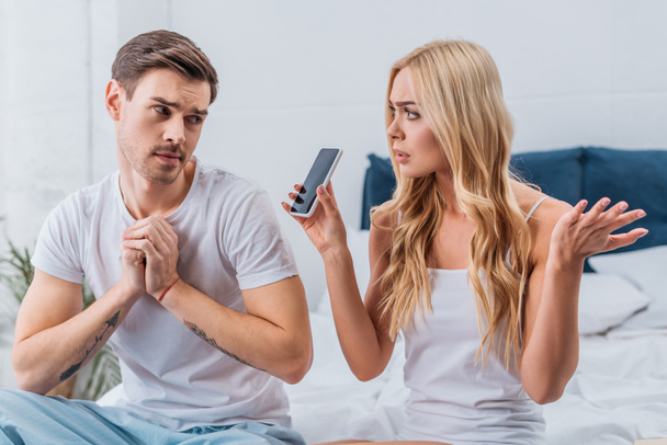 hämmentynyt nainen osoittaa älypuhelin epäilyttävä poikaystävä makuuhuoneessa
 - Valokuva, kuva