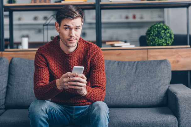 suspicious young man holding smartphone and looking at camera while sitting on couch at home - Fotoğraf, Görsel