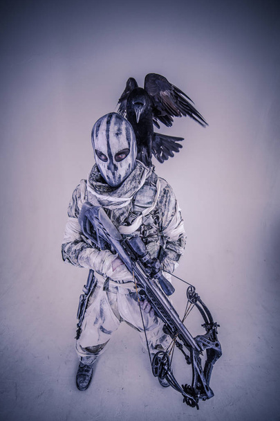 portrait of hunter man in camouflage with crossbow and raven posing against grey background - Photo, image