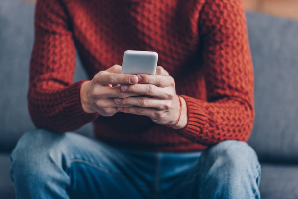 vágott lövés a fiatal ember használ smartphone otthon vörös pulóverben - Fotó, kép