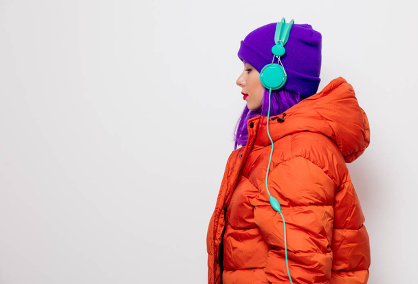 Красивая молодая девушка с фиолетовыми волосами и в оранжевой куртке слушать музыку в наушниках на белом фоне
. - Фото, изображение