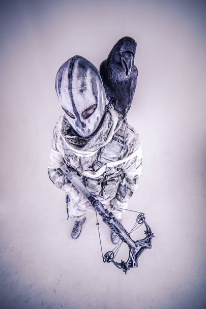 warrior man in mask with crossbow and raven posing against cold grey background - Photo, image