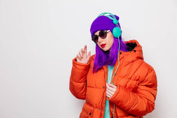 Mooi jong meisje met paarse haren en in oranje jas luisteren muziek in koptelefoon op witte achtergrond. - Foto, afbeelding