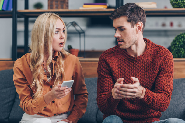 shocked young woman holding smartphone and looking at husband sitting on couch, jealousy concept - Fotó, kép