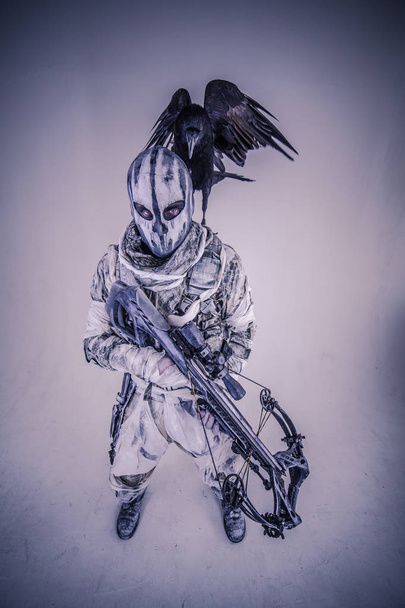 warrior man in mask with crossbow and raven posing against cold grey background - Foto, afbeelding