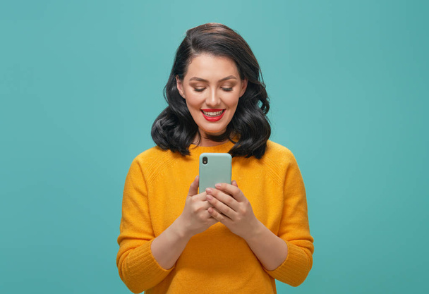 Smiling beautiful young woman in yellow sweater with mobile phone posing on turquoise background. - Φωτογραφία, εικόνα