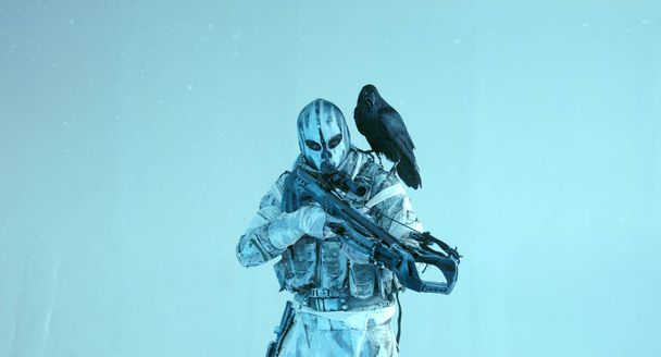 warrior in camouflage with modern crossbow and raven posing against cold blue background - Valokuva, kuva
