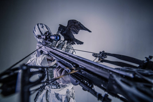 portrait of hunter man in camouflage with crossbow and raven posing against grey background - Фото, изображение