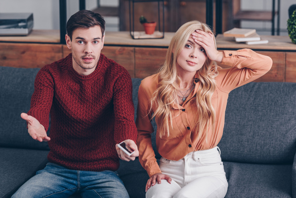 jealous man with smartphone and upset woman sitting on couch and looking at camera, distrust concept - Fotoğraf, Görsel