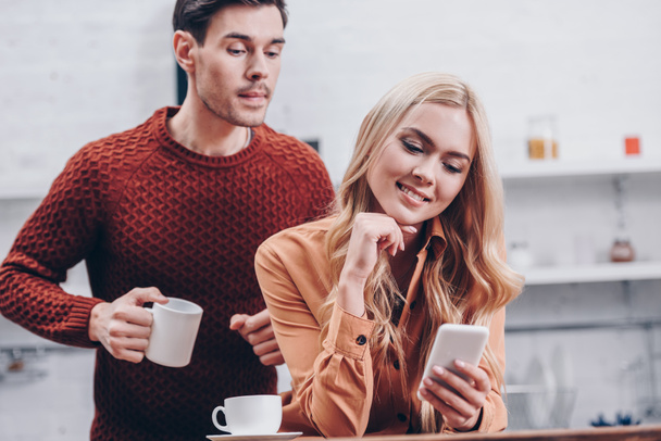 jealous young man holding cup and looking at smiling girlfriend using smartphone in kitchen, mistrust concept - Photo, image