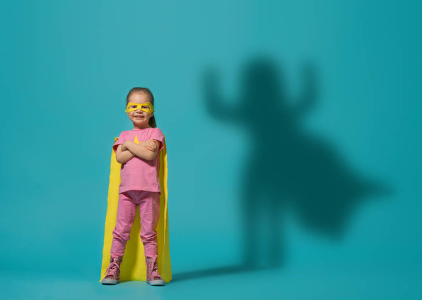スーパーヒーローを演じる小さな子供。明るい青い壁の背景に子供。女の子のパワーコンセプト。黄色、ピンク、ターコイズ色. - 写真・画像