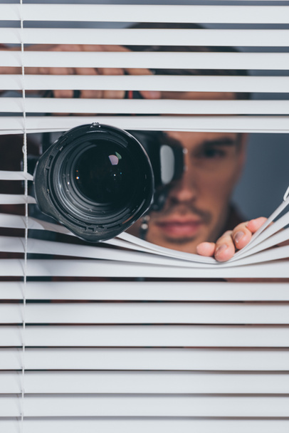 close-up view of young man holding camera and looking at camera through blinds - Photo, Image