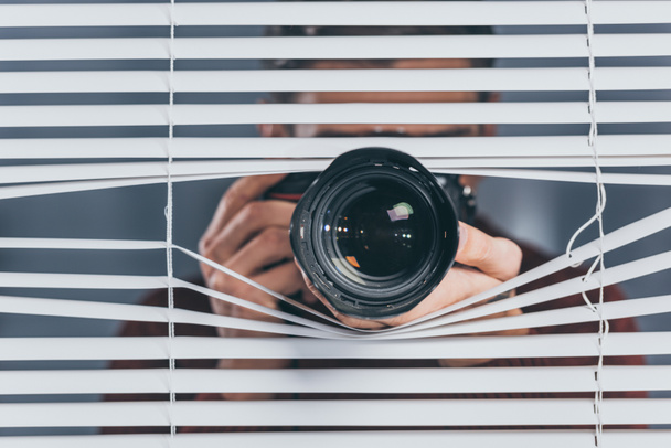 close-up view of young man holding camera and taking pictures through blinds - Photo, Image