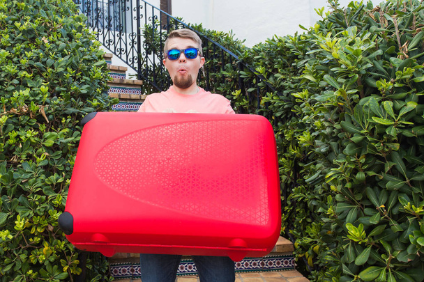 Reise-, Reise- und Urlaubskonzept - Junger Mann mit rotem Koffer in Sonnenbrille überrascht - Foto, Bild