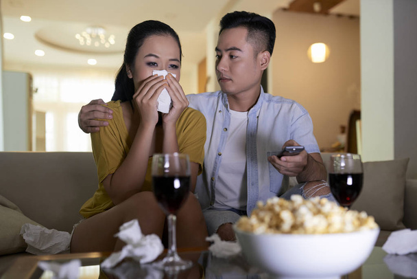 junges asiatisches Paar schaut traurigen Film und Mann tröstet weinende Frau, während sie auf Sofa sitzt - Foto, Bild