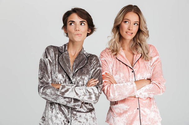 Two lovely girls wearing pajamas standing isolated over gray background, looking up - 写真・画像