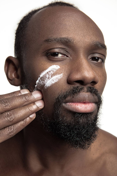 Young african-american guy applying face cream under his eyes on white background - Valokuva, kuva