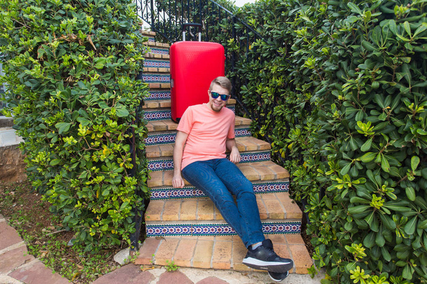 Travel, tourism and people concept - happy man sitting on stairs in sunny glasses behind the red suitcase - Фото, изображение
