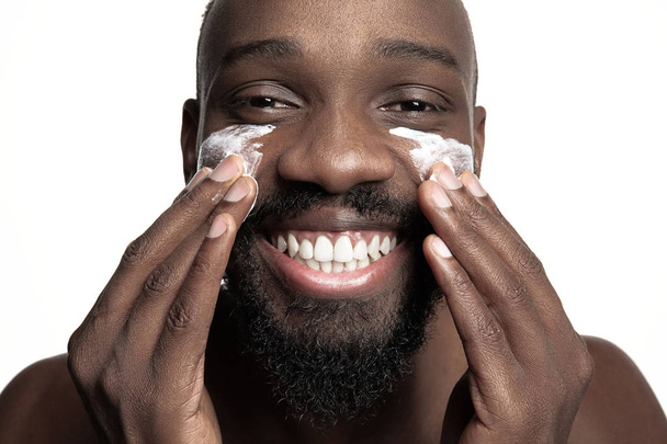 Young african-american guy applying face cream under his eyes on white background - Foto, immagini