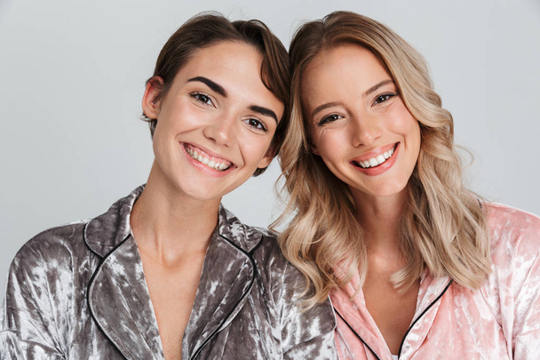 Two cheerful girls wearing pajamas standing isolated over gray background - Fotoğraf, Görsel