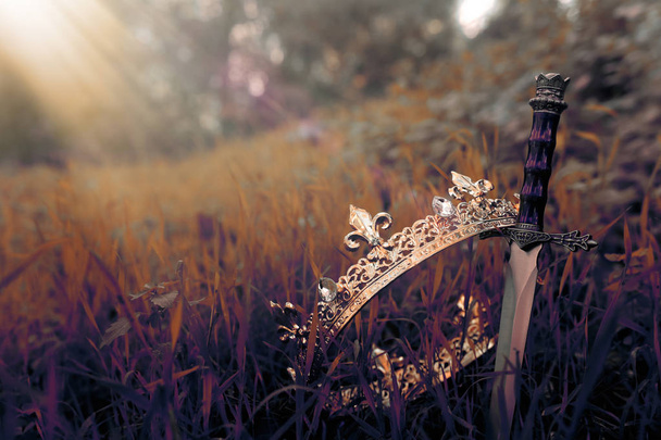 mysterious and magical photo of gold king crown and sword in the England woods or field landscape with light flare. Medieval period concept - Foto, Imagen