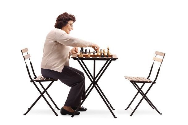 Full length profile shot of a senior woman playing chess at a table isolated on white background - Фото, изображение