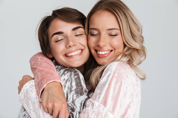 Two pretty girls wearing pajamas isolated over gray background, hugging - Zdjęcie, obraz