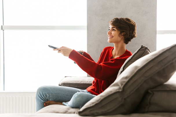 Χαμογελαστή κοπέλα φορώντας πουλόβερ, χαλαρώνοντας στον καναπέ στο σπίτι, πίνοντας καφέ, κρατώντας το τηλεχειριστήριο της τηλεόρασης - Φωτογραφία, εικόνα