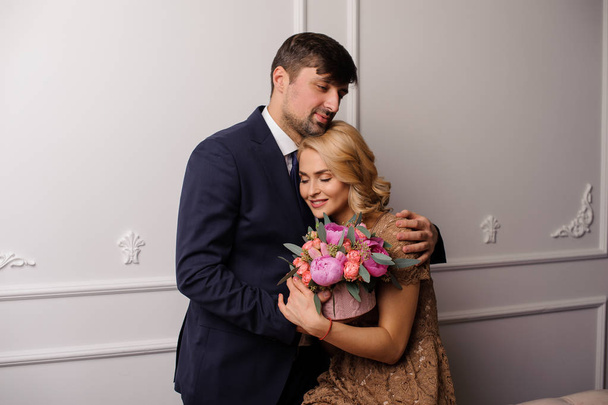 Man hugging his girl in the beige dress on the shoulder with the bouquet of flowers and looking down on the background of the white wall. - Foto, immagini