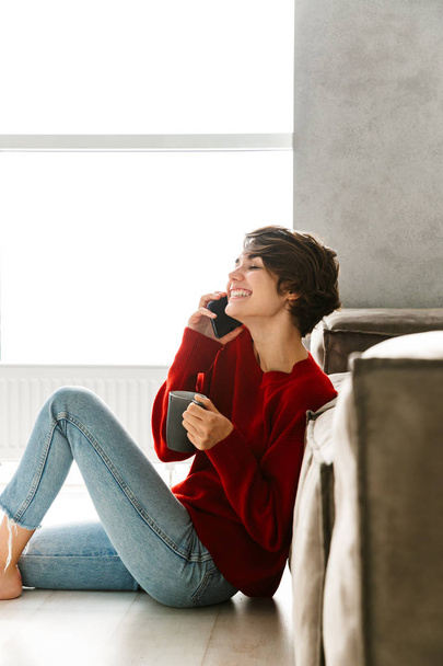 Lovely young woman wearing sweater sitting on a floor at home, drinking tea, leaning on a couch, talking on mobile phone - Фото, зображення
